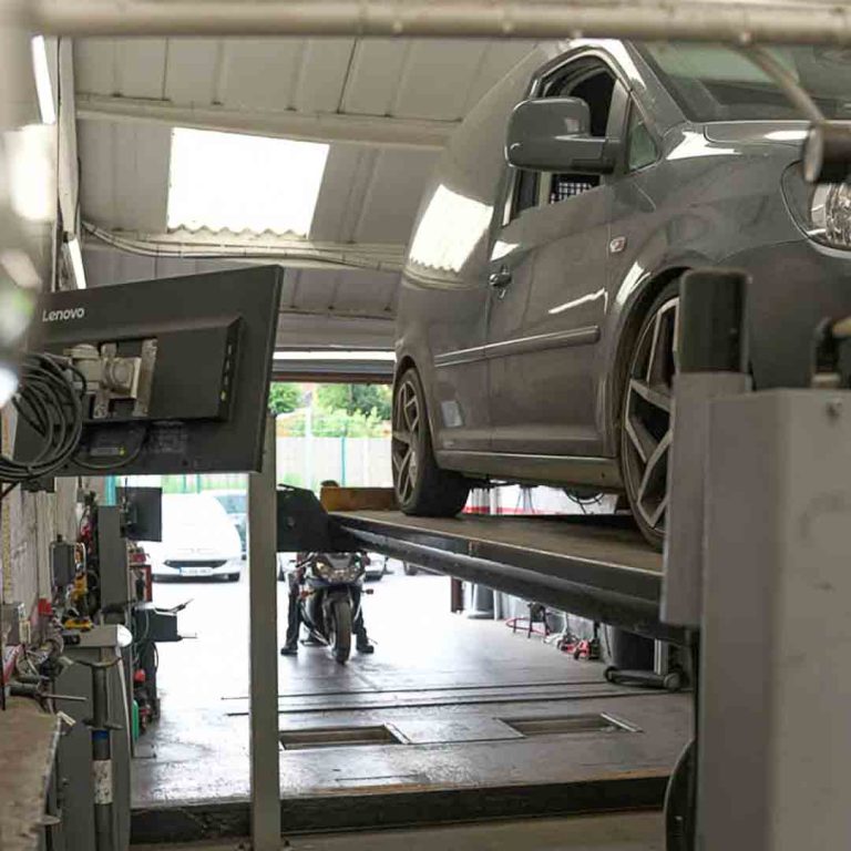 A grey car on the vehicle lift during an MOT with a bike MOT being carried out by a Motorvation Northampton technician in the background. 